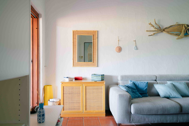 Soggiorno accogliente con divano grigio chiaro decorato con cuscini blu, un mobile in legno con frontale in vimini su cui poggiano una lampada gialla, una cesta e oggetti decorativi. Sopra il mobile è appeso uno specchio con cornice in legno e una decorazione in legno galleggiante affiancata da piccoli ornamenti sospesi. L’ambiente combina elementi naturali e moderni, creando un’atmosfera calda e invitante.